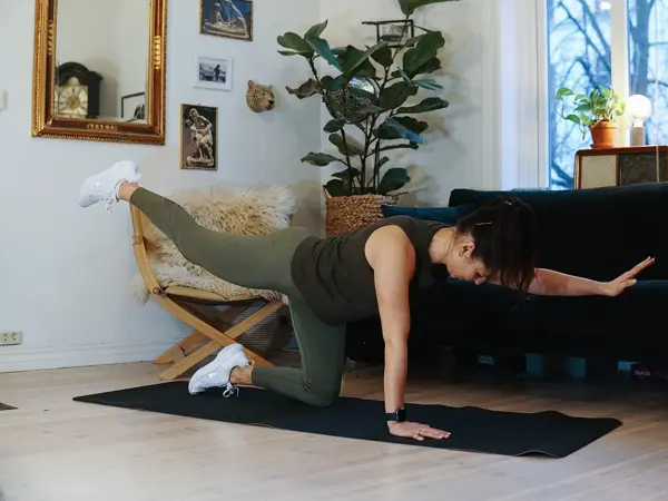 treningsekspert Anna Johansen trener på matte på gulvet, mørkegrønn sofa i bakgrunnen, lys stol med pels oppi, str grønn plante, bilder på veggen