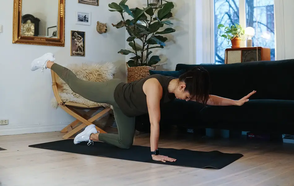 treningsekspert Anna Johansen trener på matte på gulvet, mørkegrønn sofa i bakgrunnen, lys stol med pels oppi, str grønn plante, bilder på veggen