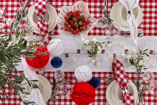 Bord dekket til fest med rødrutete duk, hvit løper, bolle med jordbær, vaser med blomster, rød, hvite og blå papirkuler, rutete servietter, beige servise.