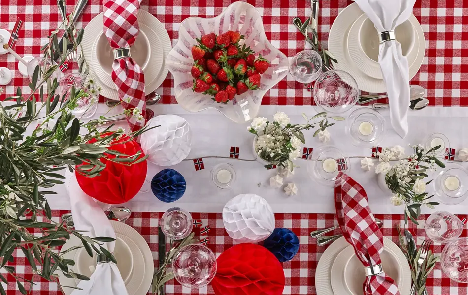 Bord dekket til fest med rødrutete duk, hvit løper, bolle med jordbær, vaser med blomster, rød, hvite og blå papirkuler, rutete servietter, beige servise.