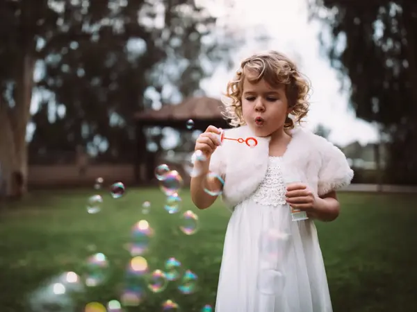 Little bridesmaid in white dress celebrating at wedding ceremony and blowing bubbles in garden