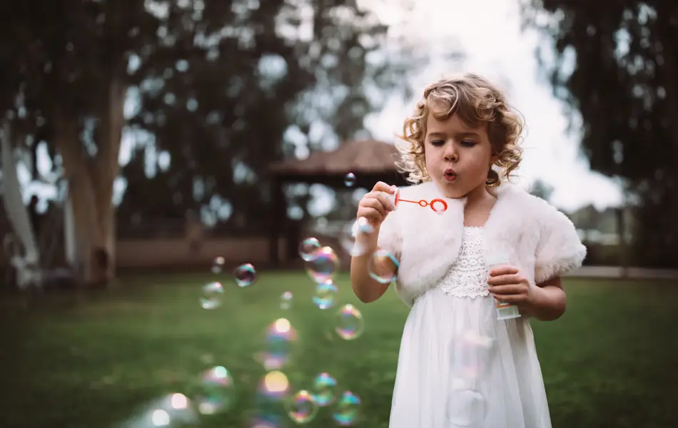 Little bridesmaid in white dress celebrating at wedding ceremony and blowing bubbles in garden