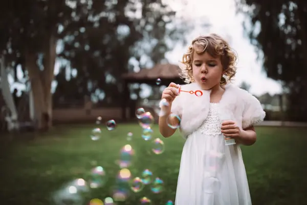 Little bridesmaid in white dress celebrating at wedding ceremony and blowing bubbles in garden