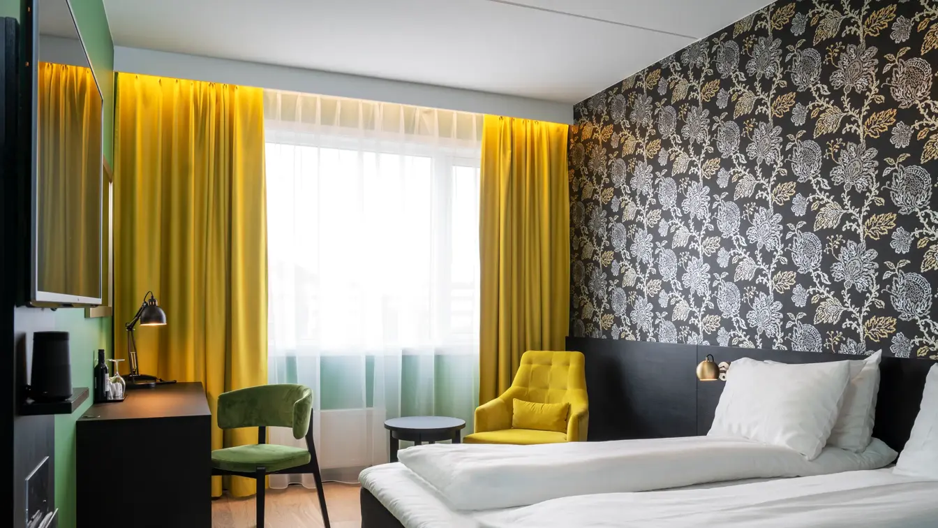 Dobbeltseng, mørk mønstrete tapet med blomster, gule gardiner, gul stol, skrivebord grønn stol og TV på dobbeltrom på Thon Hotel Kirkenes