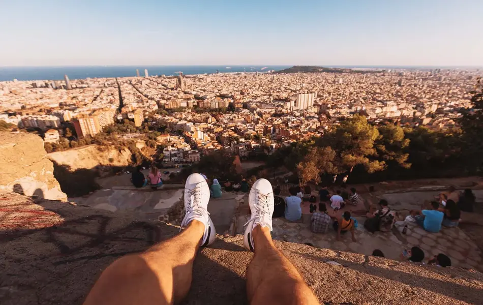 Mann sitter på en klippe. Man ser bena hans i lufta. Nedenfor er Barcelona.