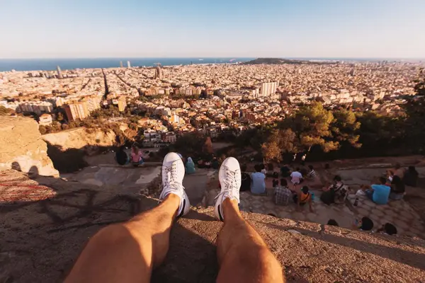 Mann sitter på en klippe. Man ser bena hans i lufta. Nedenfor er Barcelona.