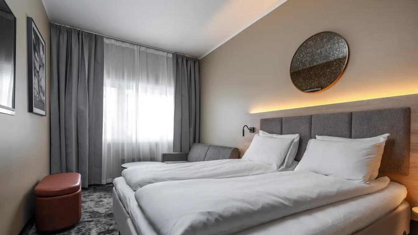 Dobbeltrom med dobbeltseng, sofa, puff, vegghengt TV, grå og hvite gardiner.