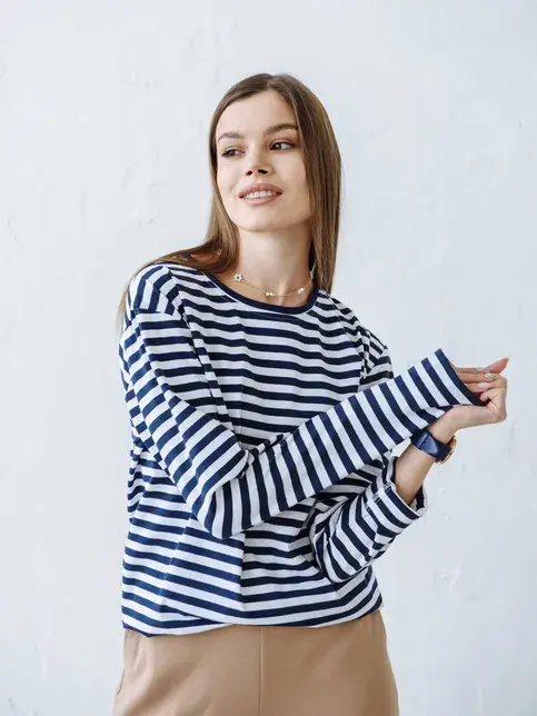 Dame med brunt, langt hår, hvit- og blåstripete genser og mørk beige bukse poserer foran kamera 