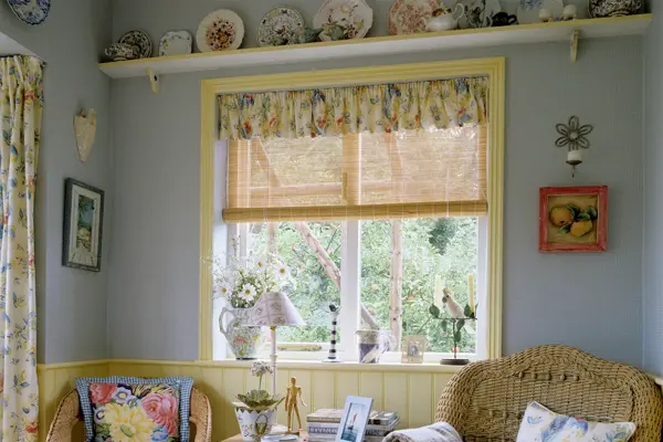 Koselig britisk interiør med to kurvstoler og et firkanta bord i et blåmalt rom, med blomstrete gardiner og vegghylle med tallerkener