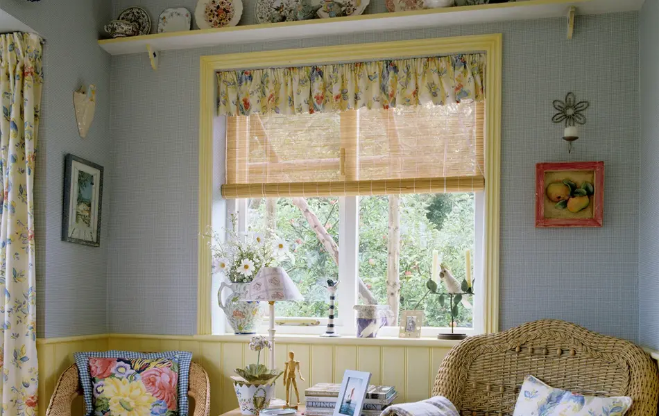 Koselig britisk interiør med to kurvstoler og et firkanta bord i et blåmalt rom, med blomstrete gardiner og vegghylle med tallerkener