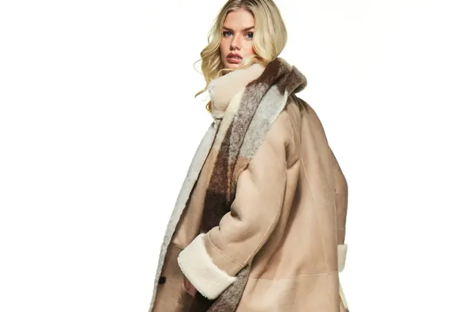 Dame snur seg mot kameraet. Hun har på seg en beige kåpe og rutet skjerf