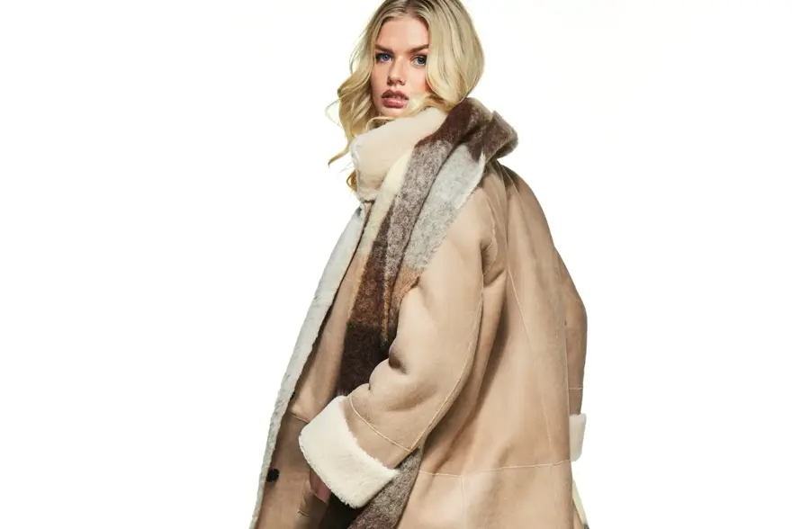 Dame snur seg mot kameraet. Hun har på seg en beige kåpe og rutet skjerf