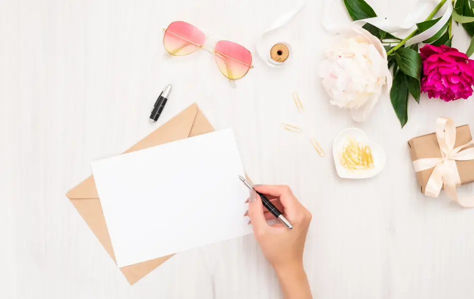 Top view woman hand writing wedding invitation card or love letter. Minimal flat lay composition with girls hand holding pen, bouquet of pion flowers, sunglasses, gift box. Writing letter concept