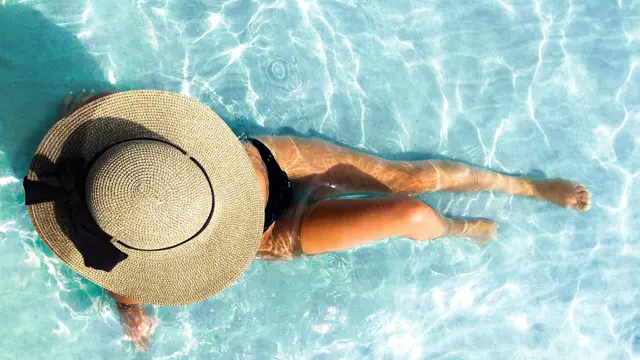 Dame ligger i blått basseng med stor rundt stråhatt 