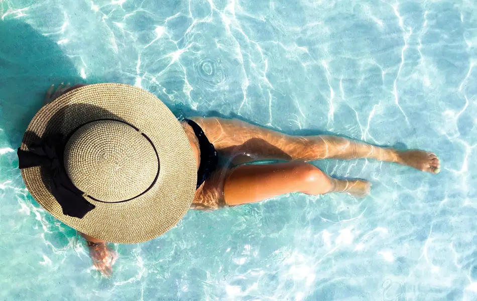 Dame ligger i blått basseng med stor rundt stråhatt 