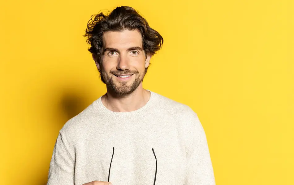 Mann med hvit ullgenser og grå ullbukse holder et par briller og poserer foran gul vegg 