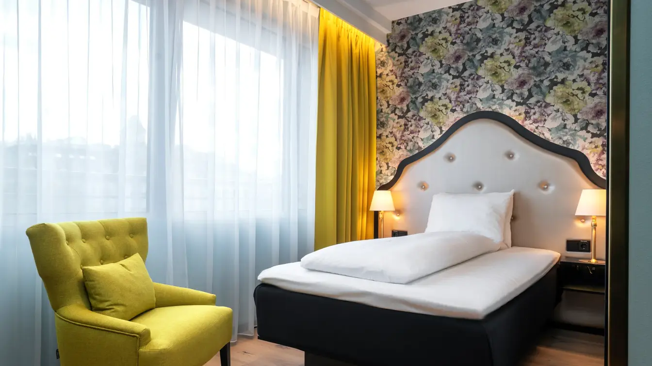 Gul stol, store vinduer, gule gardiner, blomstrete tapet og queen seng i standard rom queen på Thon Hotel Cecil.