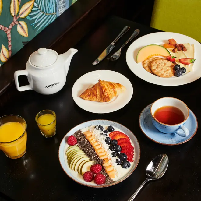 Frokostbord med tekanne, glass med juice, glass med vitaminshot, askjett med, yoghurt og frukt, frø og bær pent dandert som topping, kopp med te, askjett med croissant, askjett med kjeks, ost og frukt og skål med oppskjært frukt