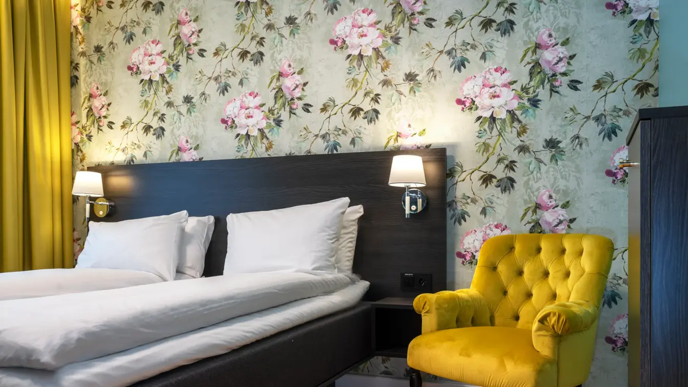 Blomstrete tapetvegg, gul stol, dobbeltseng, gule gardiner. 