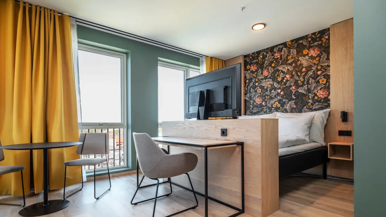 Superior rom med dobbeltseng, mønstrete tapet bak sengen, skrivebord med tilhørende stol, spiseplass, store vinduer med gule gardiner.