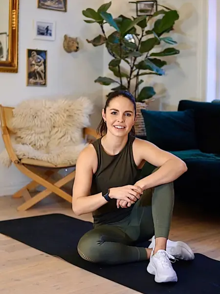 treningsekspert Anna Johansen trener på matte på gulvet, mørkegrønn sofa i bakgrunnen, lys stol med pels oppi, str grønn plante, bilder på veggen