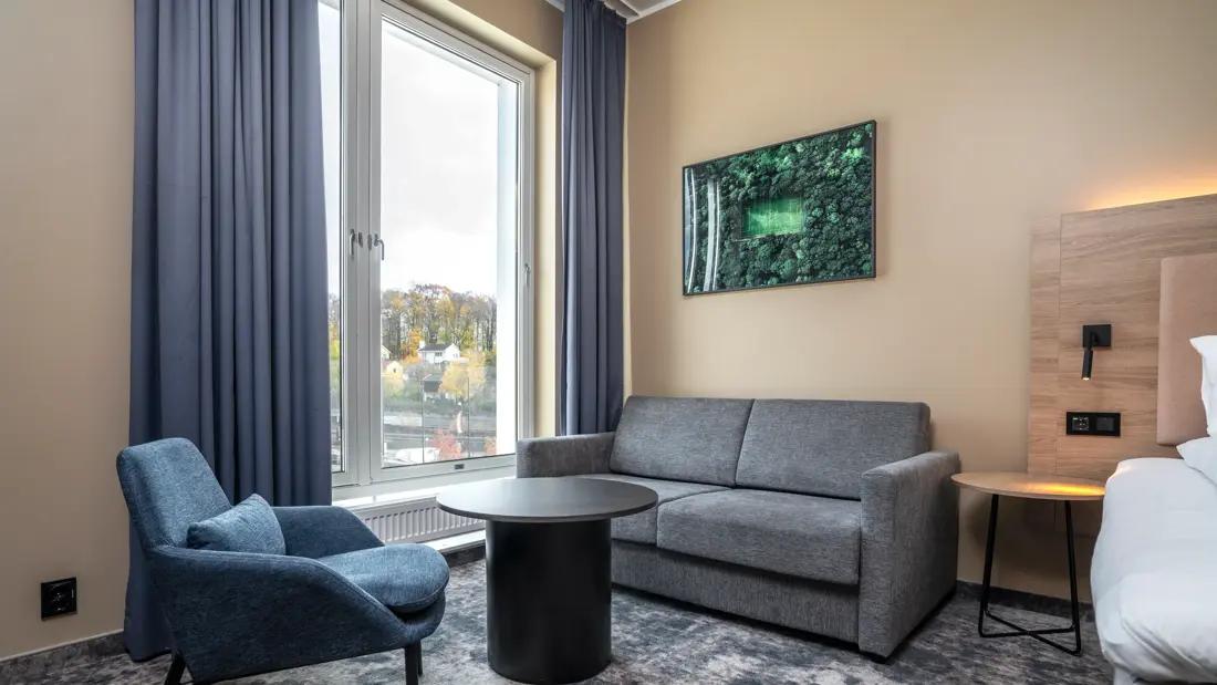 Dobbeltrom med sittegruppe, stort vindu med gardiner på hver side, vegghengt TV og teppegulv.