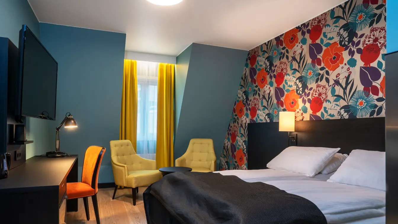 Blomstrende tapetvegg med blå vegger, gule gardiner,  vindu, to gule stoler, dobbeltseng, svart skrivebord, flatskjerm hengende på vegg. 