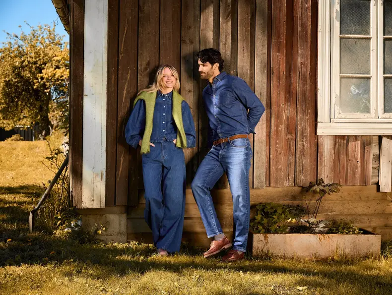 To personer i denimklær står ved en trebygning utendørs.