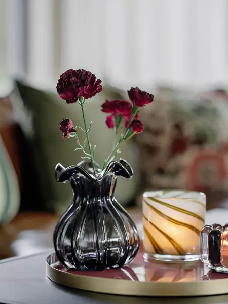 Vase med blomster og telysholdere på et brett som står på et bord. sofa med puter i bakgrunnen.