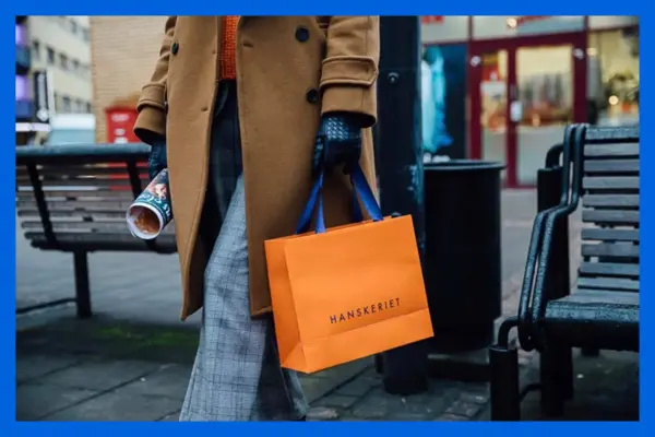 Dame i brun kåpe går ute og holder en pose fra Hanskeriet.