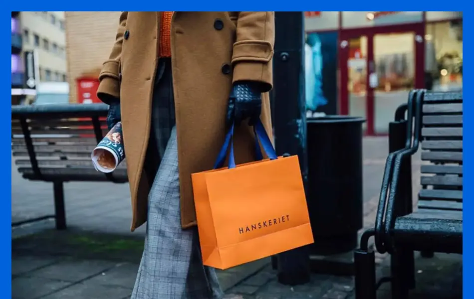 Dame i brun kåpe går ute og holder en pose fra Hanskeriet.