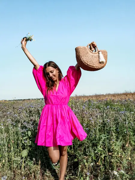 Jente i knallrosa kjole poserer med veske og blomster i blomstereng 