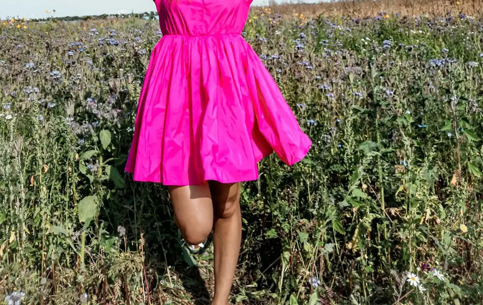 Jente i knallrosa kjole poserer med veske og blomster i blomstereng 