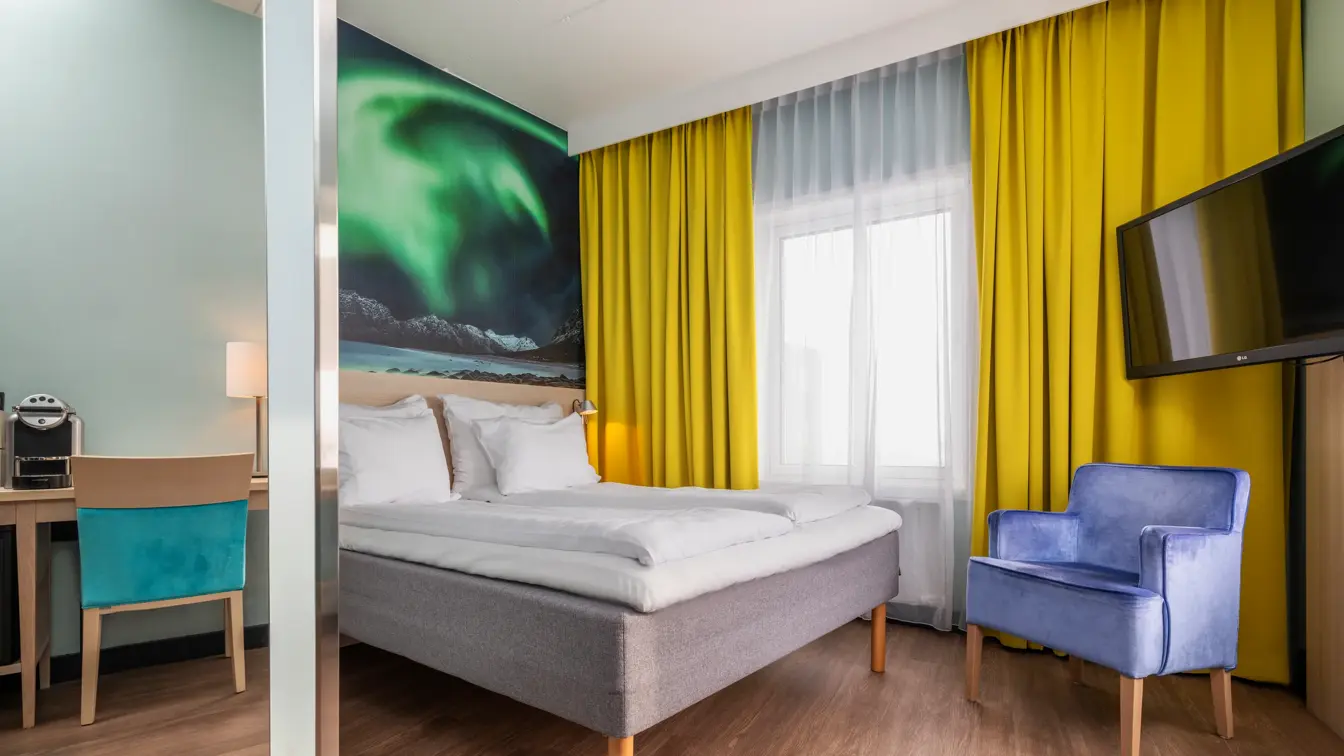 Dobbeltrom med dobbeltseng, lilla lenestol, Tv skjerm, gule gardiner og stort nordlys bilde på veggen.