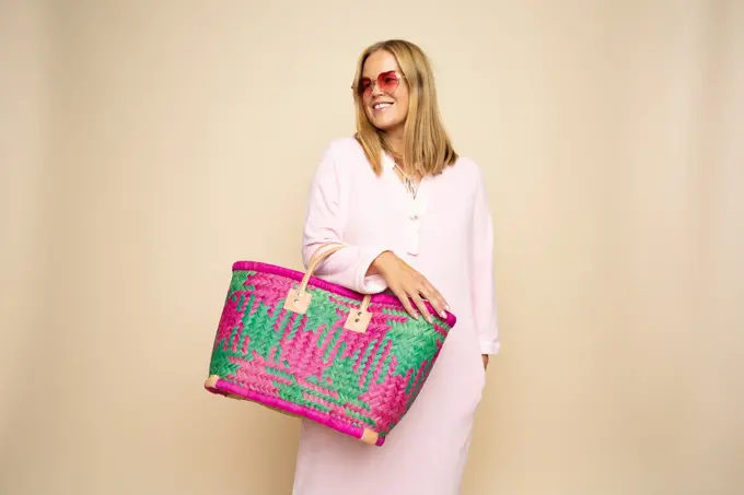 Damemodell i lyserosa lang kjole, rosa solbriller, stor flettet rosamønstret stråveske,