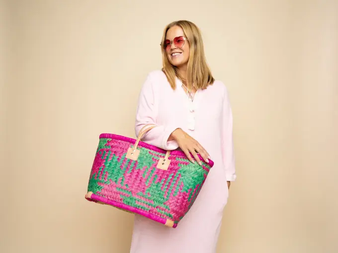 Damemodell i lyserosa lang kjole, rosa solbriller, stor flettet rosamønstret stråveske,