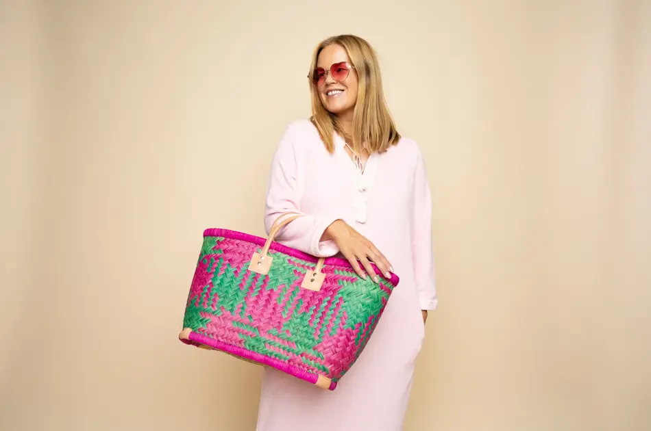 Damemodell i lyserosa lang kjole, rosa solbriller, stor flettet rosamønstret stråveske,