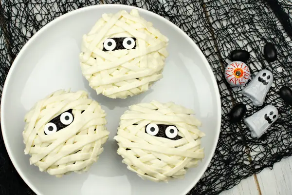 Group of Halloween mummy cupcakes on white plate with candies on black cloth background