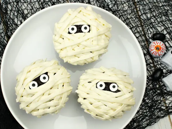 Group of Halloween mummy cupcakes on white plate with candies on black cloth background