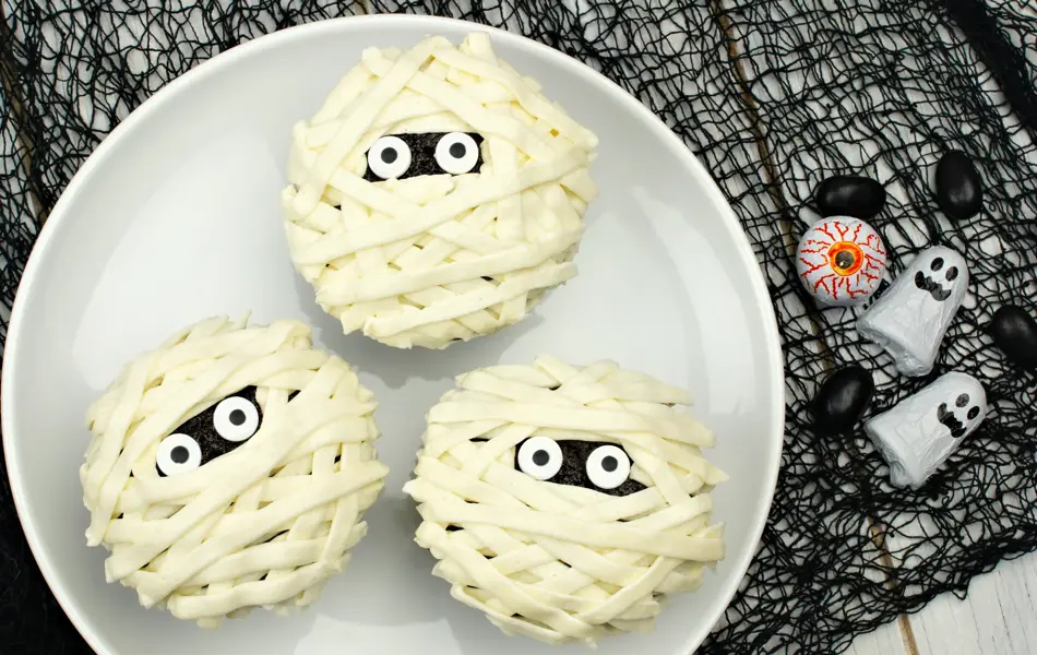 Group of Halloween mummy cupcakes on white plate with candies on black cloth background
