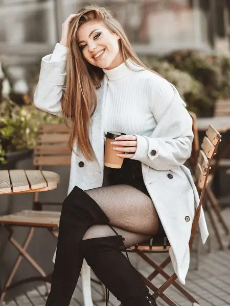 Jente med svarte, knelange støvletter og grå kåpe sitter på stol og smiler til kamera