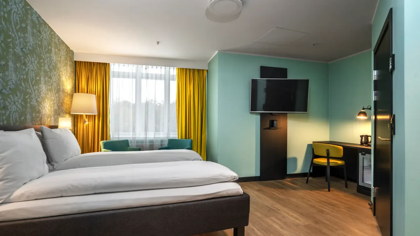 Dobbeltseng, gull gardiner i bakgrunnen, TV hengt opp på veggen og skrivebord med gul stol i høyre hjørne. 