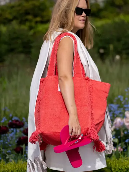 Dame står ute med hvit kjole, et håndkle over skulderen, en stor rød stoffveske over skulderen og holder et par rosa flip flops