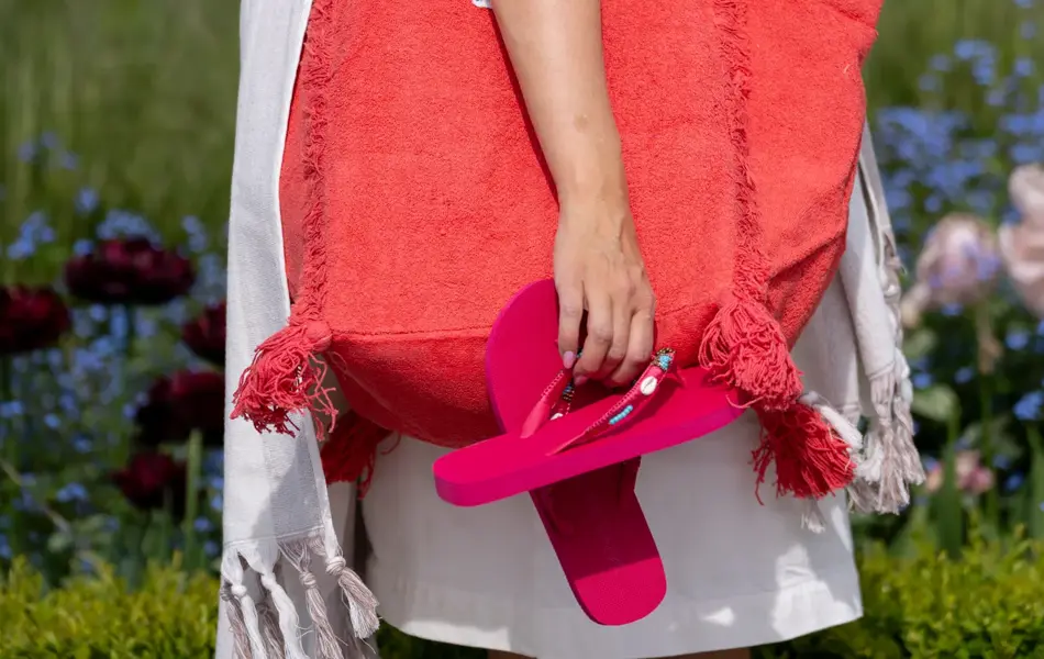 Dame står ute med hvit kjole, et håndkle over skulderen, en stor rød stoffveske over skulderen og holder et par rosa flip flops