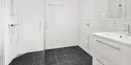 Tomt bad med hvit fliser på vegg og mørke fliser på gulv, dusjhjørne med glassvegger