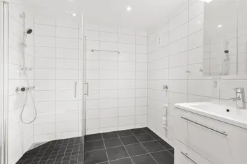 Tomt bad med hvit fliser på vegg og mørke fliser på gulv, dusjhjørne med glassvegger