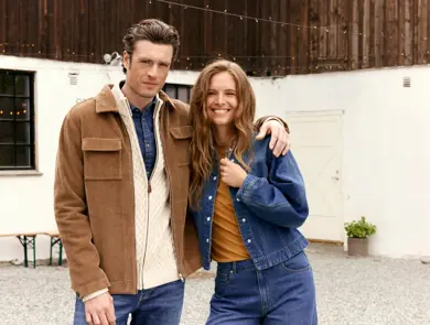 En mann i beige cardigan og brun jakke holder rundt en kvinne i brun topp og jeansjakke.