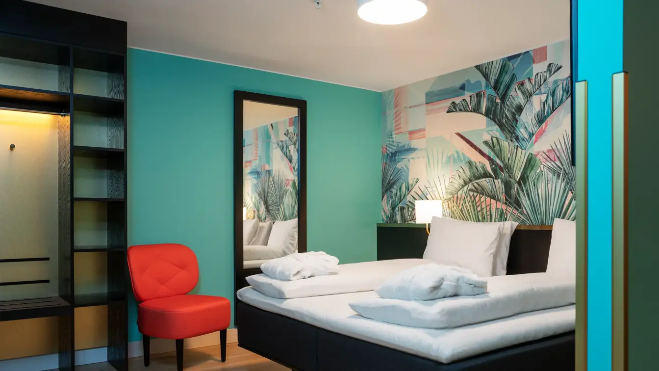 Fargerik tapet med jungelblad, dobbeltseng med morgenkåper, turkis vegg med stort speil og en rød stol i junior suite på Thon Hotel Storo.