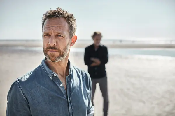 Eldre mann i olaskjorte på strand. To menn. Mann i bakgrunnen.