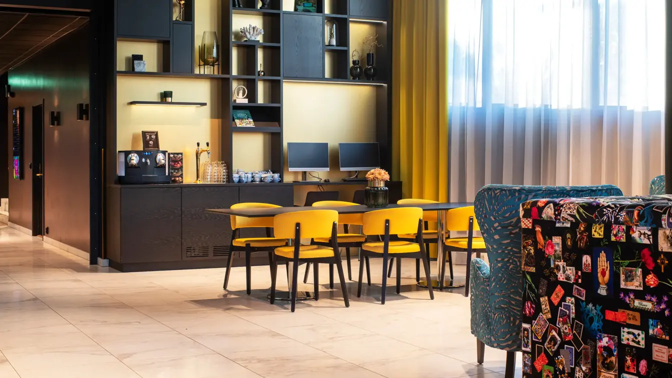 Sort hyllseksjon med kaffemaskin og PCer, sort bord med gule stoler og gule gardiner i resepsjonen på Thon Hotel Storo